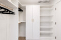 Bright white walk-in closet with open shelving on the right, hanging rod with black hangers on the left, and a tall central cabinet with gold handles.