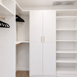 Bright white walk-in closet with open shelving on the right, hanging rod with black hangers on the left, and a tall central cabinet with gold handles.