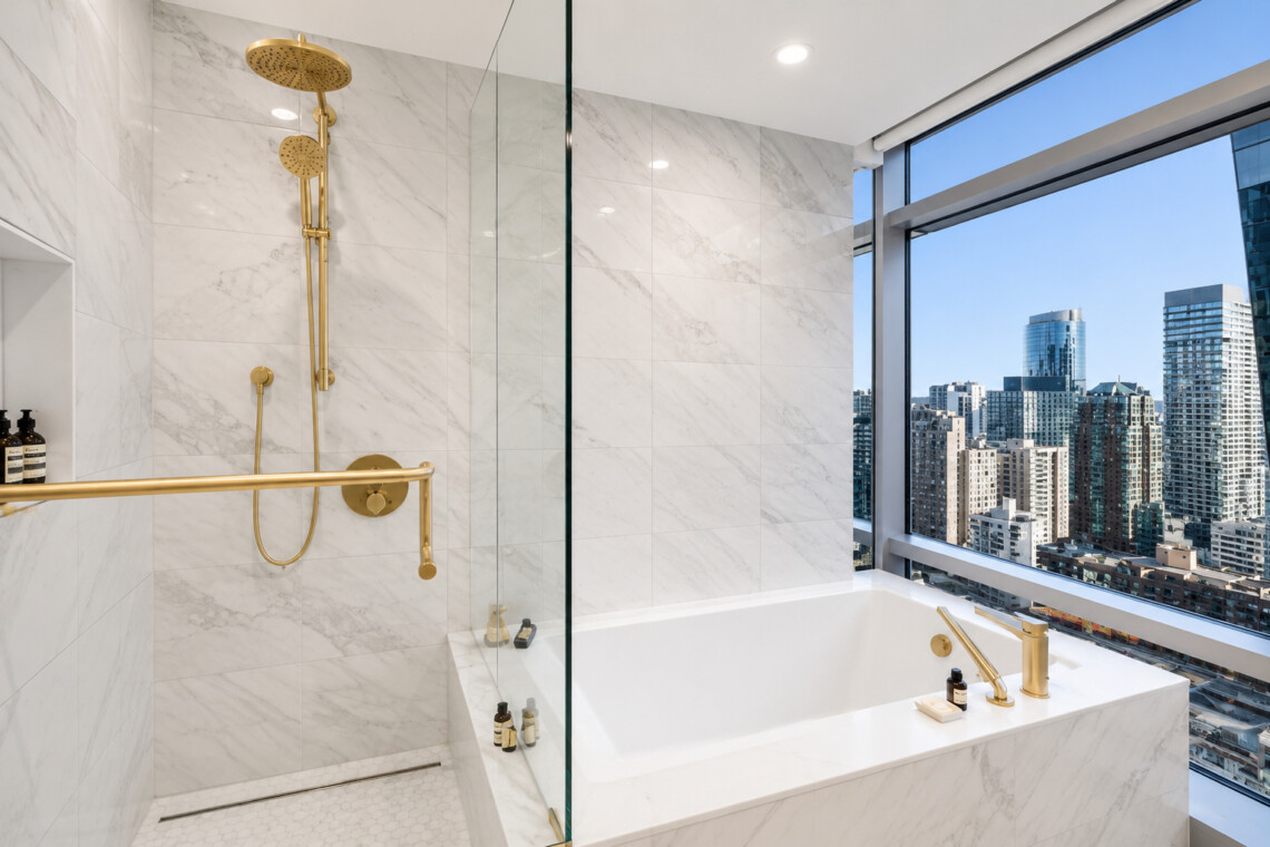 Luxurious marble bathroom with gold fixtures, a glass shower, and a city view through a large window at the tub area.