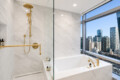 Luxurious marble bathroom with gold fixtures, a glass shower, and a city view through a large window at the tub area.