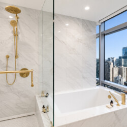 Luxurious marble bathroom with gold fixtures, a glass shower, and a city view through a large window at the tub area.