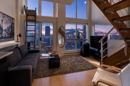 Loft living room with large windows showing a city skyline at sunset, wooden stairs, and a gray sofa on a shag rug.