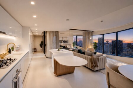Open-plan kitchen and living area in a high-rise apartment, featuring a curved white island, beige sofa, and city skyline at sunset.