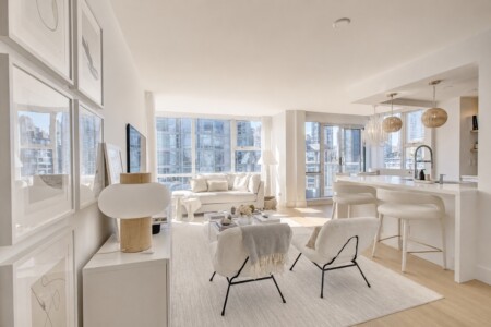 Bright, minimalist open-plan living area with white furniture, a beige sofa, two armchairs, and a kitchen island with white stools; city buildings visible through large windows.
