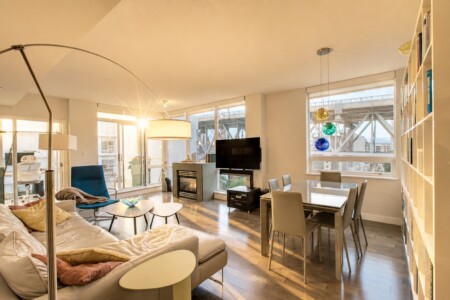 Bright open-concept living room with a fireplace, TV, and glass dining table by large windows overlooking a cityscape.