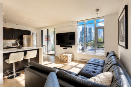 Open-plan living room with a black leather sofa, tufted ottoman, wall-mounted TV, and large windows overlooking a city skyline.