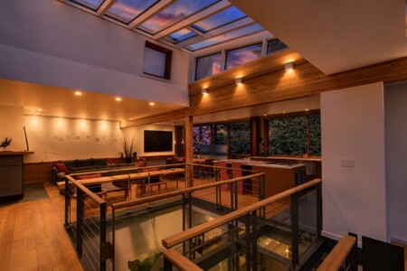 Bright, modern open-concept living area with skylit ceiling, wooden beams, and a glass floor railing over a sunken lower level.