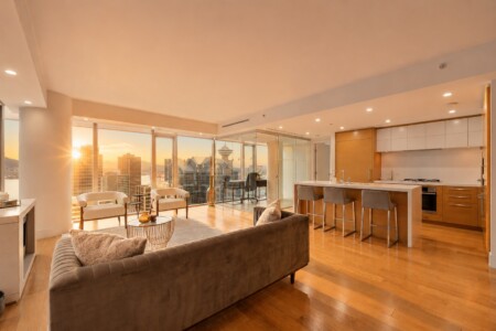 Sunlit open-concept living room and kitchen in a high-rise apartment with floor-to-ceiling windows overlooking the city skyline at sunset.