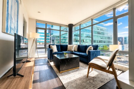 Bright, modern living room with a navy blue sectional sofa, cream cushions, and a black marble coffee table, all bathed in natural light from large glass walls framing a city view.