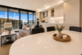 Open-plan living area with a white oval dining table, candles on a woven tray, gray sofa, and floor-to-ceiling windows overlooking sailboats at a marina.