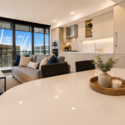 Open-plan living area with a white oval dining table, candles on a woven tray, gray sofa, and floor-to-ceiling windows overlooking sailboats at a marina.