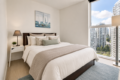 Cozy modern bedroom with a queen bed, white linens, gray throw, and a large window showing city high-rises outside.