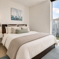 Cozy modern bedroom with a queen bed, white linens, gray throw, and a large window showing city high-rises outside.