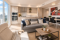 Open-concept living area with a gray sofa, beige chair, coffee table, and a dining area; large windows reveal city skyline on a sunny day.