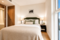 A cozy hotel‑style bedroom with a queen bed, green headboard, white linens, beige throw, and two matching nightstands with lamps flanking the bed; a framed art print hangs above, and a large window shows a city view.