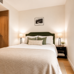 A cozy hotel‑style bedroom with a queen bed, green headboard, white linens, beige throw, and two matching nightstands with lamps flanking the bed; a framed art print hangs above, and a large window shows a city view.