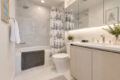 Modern bathroom with white bathtub, gray towel, patterned shower curtain, toilet, and light wood vanity with a large backlit mirror edge.
