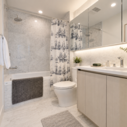 Modern bathroom with white bathtub, gray towel, patterned shower curtain, toilet, and light wood vanity with a large backlit mirror edge.