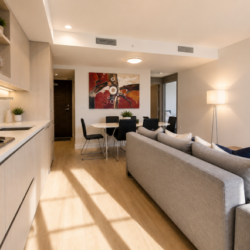 Open-concept living area with a light wood kitchen on the left, a gray sofa and a wall-mounted TV on the right, and a dining table in the back.