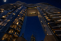 Night view of a modern curved glass building connected by a skybridge, with illuminated windows and a full moon in a starry sky.
