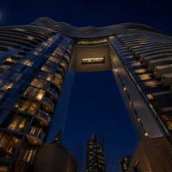Night view of a modern curved glass building connected by a skybridge, with illuminated windows and a full moon in a starry sky.