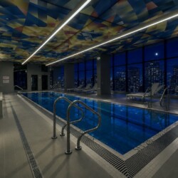 Indoor pool area in a modern gym at night, with a colorful patterned ceiling, bright linear lights, and city lights visible through large windows.