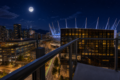Night city view from a balcony: glowing streets, glass buildings, and a lit stadium with a distinctive roof under a full moon.