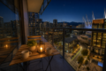 Nighttime balcony with a wooden table set for two: two mugs, a small plant, and a lantern, overlooking a lit cityscape.