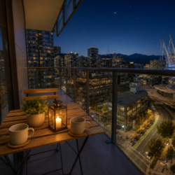 Nighttime balcony with a wooden table set for two: two mugs, a small plant, and a lantern, overlooking a lit cityscape.