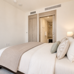 Hotel bedroom with a large bed, beige bedding and pillows; open doorway reveals an en suite bathroom in the background.