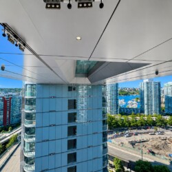 High-angle view of a modern city skyline with glass towers, a marina with boats, and a construction site below, seen from beneath a large overhang.