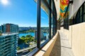 Sunlit modern hallway with large windows overlooking a river and city, colorful geometric ceiling artwork overhead and bright blue sky outside.