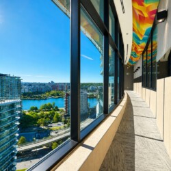 Sunlit modern hallway with large windows overlooking a river and city, colorful geometric ceiling artwork overhead and bright blue sky outside.