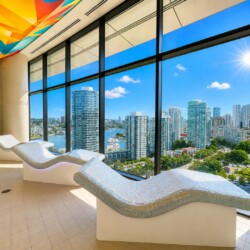 Indoor wellness lounge with curved mosaic-tile loungers facing large windows overlooking a sunny city skyline and marina.