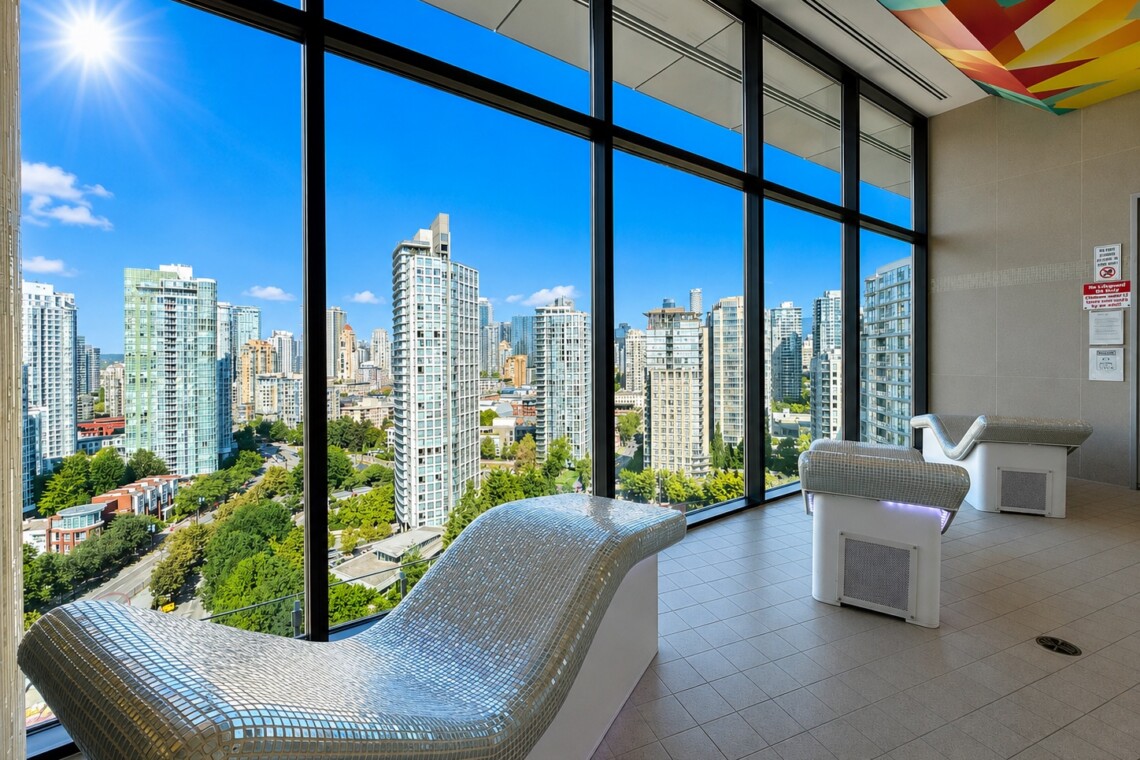 Indoor spa lounge with two curved mosaic-tile chaise lounges facing floor-to-ceiling windows overlooking a sunny city skyline.