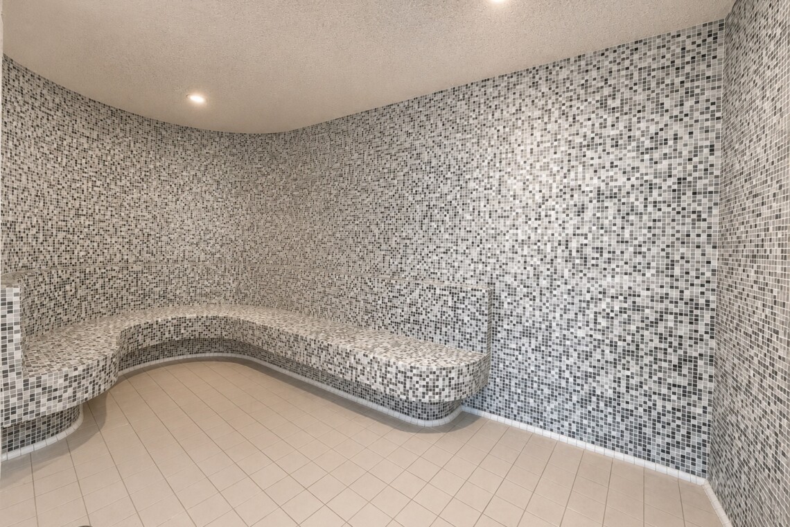 Curved mosaic-tiled steam room with built-in bench along the walls, illuminated by recessed ceiling lights.