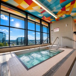 Indoor hot tub by large windows with a city view; colorful geometric ceiling above and stainless handrails nearby.