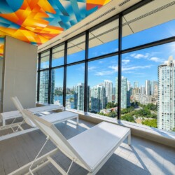 Indoor lounge area with white sunbeds and a bright multi-colored geometric ceiling, overlooking a city skyline through floor-to-ceiling windows