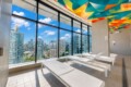 Indoor pool deck with white lounge chairs and large glass windows overlooking a city skyline; colorful geometric ceiling art above.