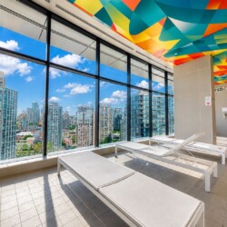 Indoor pool deck with white lounge chairs and large glass windows overlooking a city skyline; colorful geometric ceiling art above.