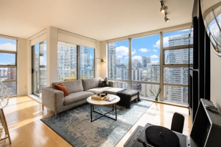 Bright city-view living room with large windows, grey sectional sofa, and oval coffee table on a blue rug.