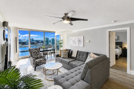 Bright modern living room with a gray sectional sofa, patterned armchair, and round marble coffee table, facing large windows with a river and city skyline outside.