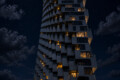 Tall modern apartment tower with staggered balconies, illuminated windows, against a starry night sky and a full moon nearby.
