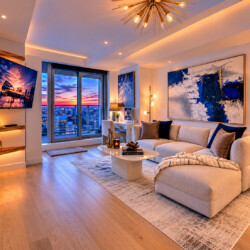 Contemporary living room with a beige sectional, glass coffee table, and bold blue abstract art, overlooking a city skyline at sunset from large windows.