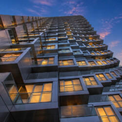 Low-angle view of a high-rise with stacked glass balconies glowing at sunset.