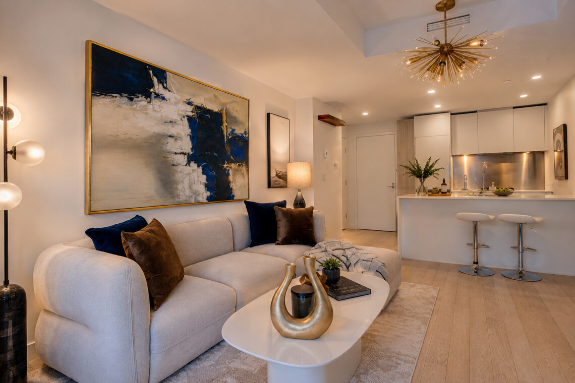 Open-plan living room with a beige sofa, blue and brown cushions, abstract wall art, and a white coffee table with a gold sculpture.