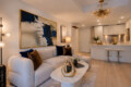 Open-plan living room with a beige sofa, blue and brown cushions, abstract wall art, and a white coffee table with a gold sculpture.
