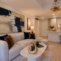 Open-plan living room with a beige sofa, blue and brown cushions, abstract wall art, and a white coffee table with a gold sculpture.