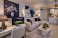 Modern living room with a light gray sectional, abstract wall art, and a marble desk near a monitor; kitchen visible in the background.