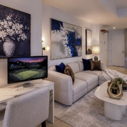 Modern living room with a light gray sectional, abstract wall art, and a marble desk near a monitor; kitchen visible in the background.